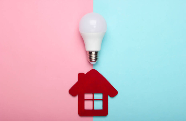 Energy saving concept. House figurine and energy-saving light bulb on a blue-pink pastel background. Top view - 写真・画像
