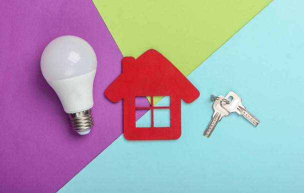 Energy saving concept. House figurine with keys and energy-saving light bulb on colored background. Top view - Zdjęcie, obraz