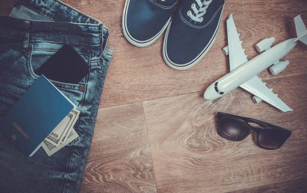 Concepto de viaje o turismo. Jeans, billetes de cien dólares, zapatillas, smartphone, pasaporte, gafas de sol, avión en un piso de madera. Vista superior. Puesta plana - Foto, Imagen