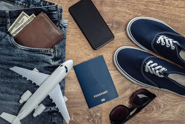 Concepto de viaje o turismo. Jeans, billetes de cien dólares, zapatillas, smartphone, pasaporte, gafas de sol, avión en un piso de madera. Vista superior. Puesta plana - Foto, imagen