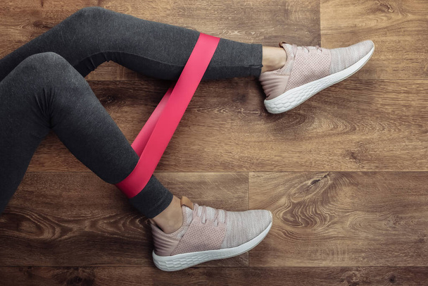 Gambe femminili in leggings sportivi e scarpe da ginnastica che fanno esercizi con elastico d'idoneità su un piano di legno. Allenamento fitness a casa. Vista dall'alto - Foto, immagini