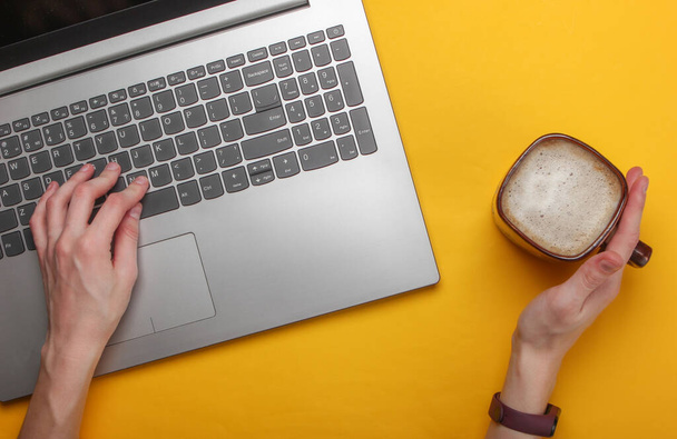 Kadın eli bir bardak kapuçino tutuyor ve sarı arka planda dizüstü bilgisayar kullanıyor. Serbest konsept, çalışma alanı, online çalışma. Üst görünüm - Fotoğraf, Görsel