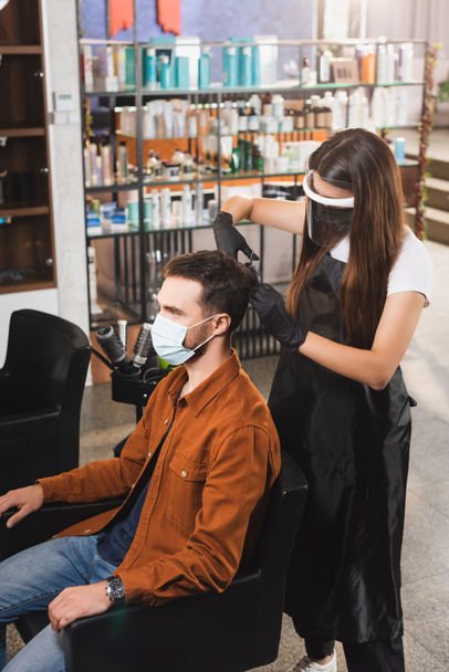 hairstylist in face shield and latex gloves cutting hair of client wearing medical mask - Fotoğraf, Görsel