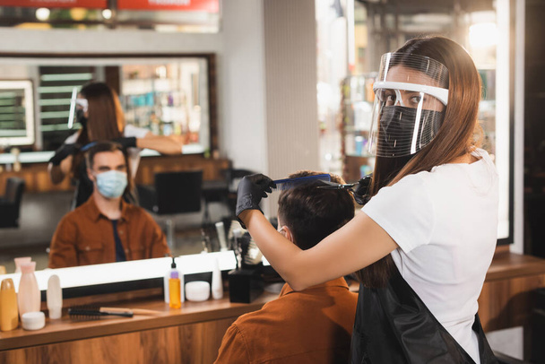 парикмахер в лице щит стрижет волосы клиента в медицинской маске рядом с размытым зеркальным отражением - Фото, изображение
