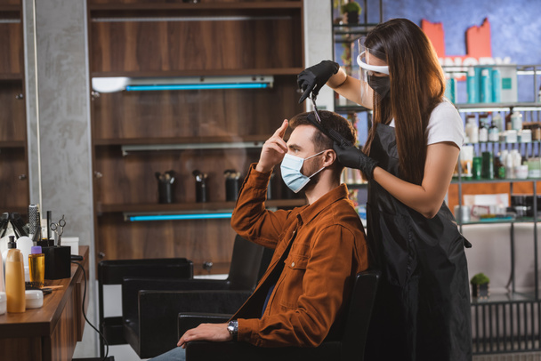 side view of man pointing with fingers at his hair while hairdresser in face shield making haircut - 写真・画像