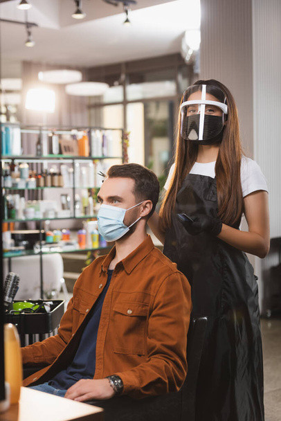 coiffeur dans le bouclier facial regardant la caméra tout en se tenant près du client portant un masque médical - Photo, image