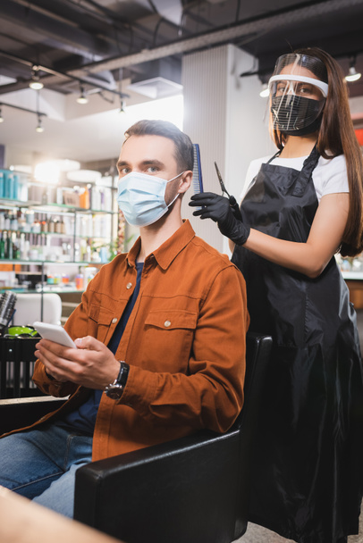 hairdresser in protective equipment looking at camera near client holding smartphone - Photo, Image