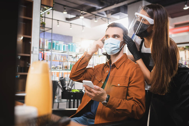 mężczyzna w masce medycznej trzymając smartfona i wskazując palcem blisko fryzjera w tarczy twarzy, baner - Zdjęcie, obraz