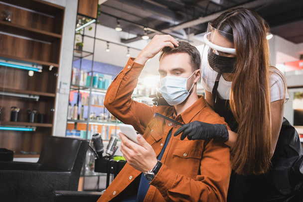 barber in face shield and latex gloves near client touching his hair and holding smartphone - Foto, Bild