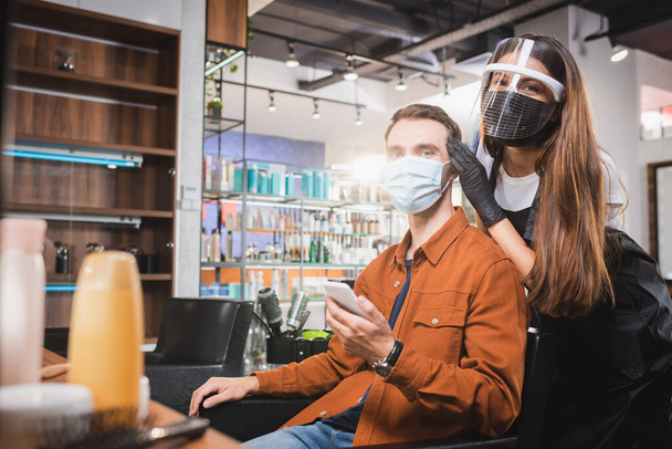 cabeleireiro no escudo facial olhando para a câmera perto do homem em máscara médica segurando smartphone, primeiro plano borrado - Foto, Imagem