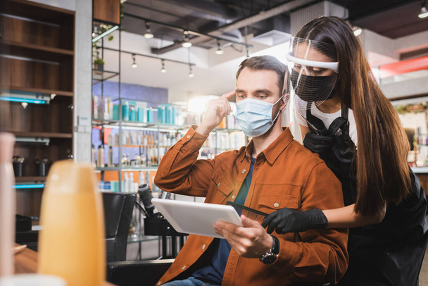 hombre con máscara médica sosteniendo la tableta y apuntando con el dedo a su cabello cerca de peluquería sosteniendo peine - Foto, imagen
