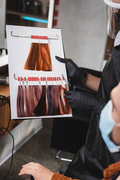 vista recortada del peluquero en guantes de goma que sostienen la paleta de colores de pelo cerca del cliente en primer plano borroso - Foto, Imagen