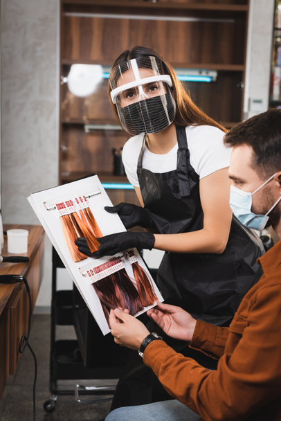 Friseur im Gesichtsschutz schaut in die Kamera, während er dem Kunden Haarfarben zeigt - Foto, Bild