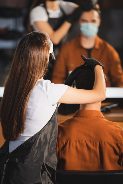 Friseur in Schürze und Latexhandschuhen berührt Haare des Mannes in medizinischer Maske in der Nähe verschwommener Spiegelreflexion - Foto, Bild