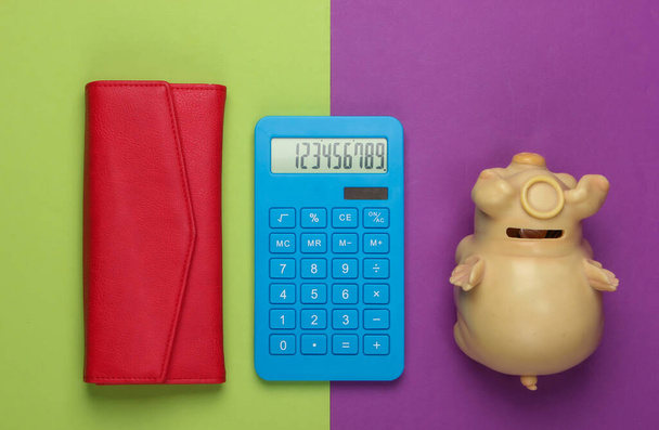Gerenciar orçamento familiar. Custos de compras. Calculadora azul com carteira de couro vermelho e mealheiro no fundo verde roxo. Vista superior - Foto, Imagem