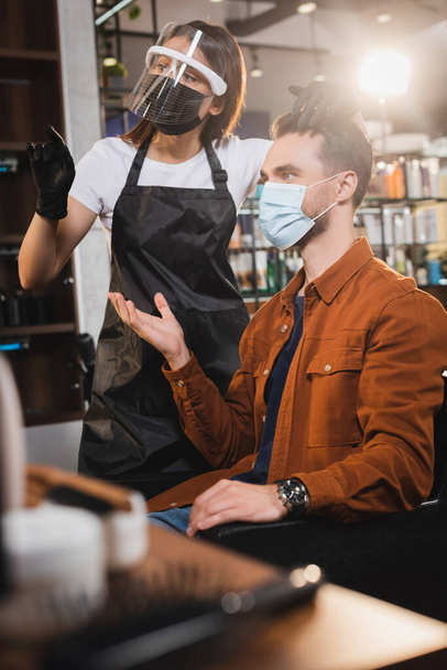 hairstylist in face shield and latex gloves pointing with finger, and client in medical mask pointing with hand, blurred foreground - Fotoğraf, Görsel