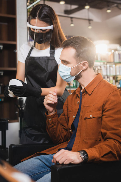 barbier dans le bouclier facial contenant tenant avec baume pour les cheveux près du client dans le masque médical - Photo, image