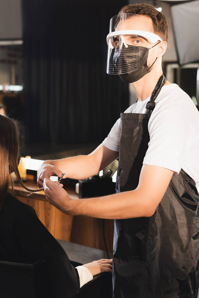peluquero en el escudo facial cortar el pelo del cliente mientras mira a la cámara - Foto, Imagen