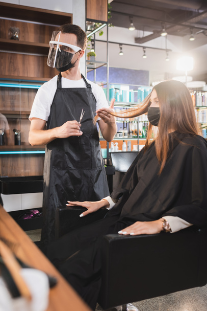 hairdresser in face shield holding scissors and touching hair of client, blurred foreground - Photo, Image