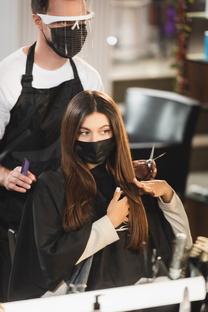 femme en masque médical touchant ses cheveux près du coiffeur dans le bouclier facial tenant des ciseaux et peigne - Photo, image