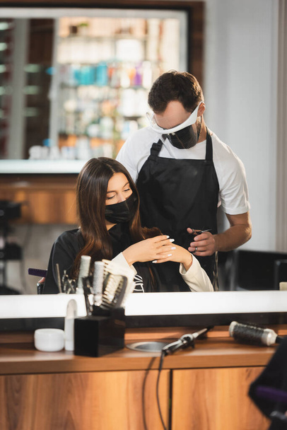 reflejo espejo de peluquería en el escudo facial cerca de la mujer en máscara médica con herramientas de peluquería en primer plano borrosa - Foto, imagen