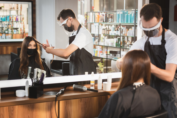 дзеркальне відображення перукаря, що ріже волосся клієнта в медичній масці, розмитий передній план
 - Фото, зображення