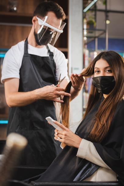 жінка в медичній масці, використовуючи смартфон, в той час як перукар в обличчя щит, що ріже волосся, розмитий передній план
 - Фото, зображення