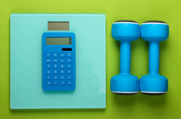 Fitness, weight loss still life. Calorie Counting. Calculator, dumbbells and scales on green background. Minimalism. Flat lay. Top view - Photo, Image