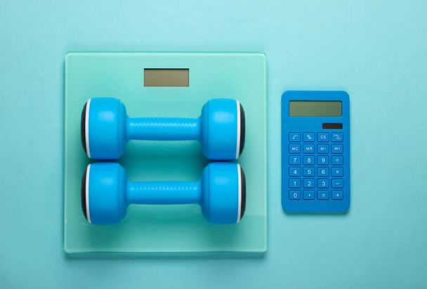 Fitness, weight loss still life. Calorie Counting. Calculator, dumbbells and scales on blue background. Minimalism. Flat lay. Top view - Photo, Image