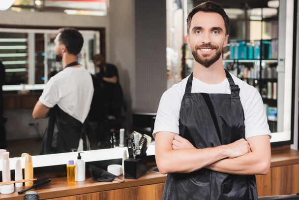 cheerful hairdresser looking at camera while standing with crossed arms on blurred background - Photo, Image