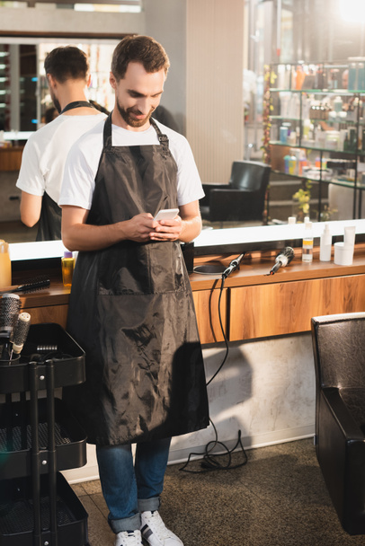 jovem barbeiro conversando no telefone celular no local de trabalho perto do espelho - Foto, Imagem