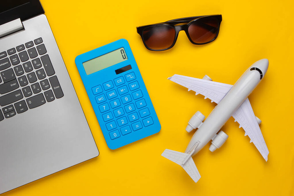 Cálculo do custo do turismo ou emigração. Reserva online. Laptop, avião, calculadora e óculos de sol em um fundo amarelo. Vista superior - Foto, Imagem