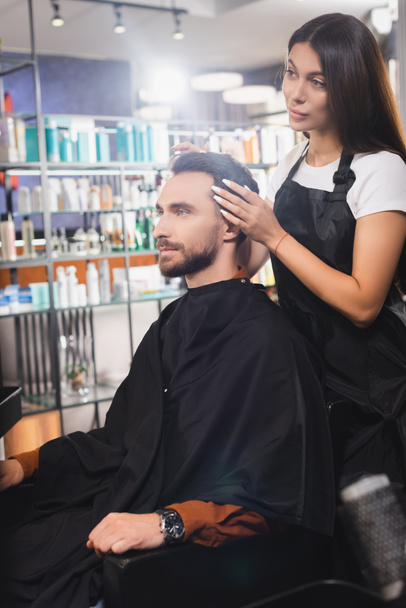 barber in apron touching hair of young bearded man in barbershop, blurred foreground - Фото, изображение
