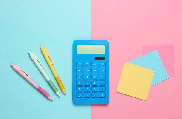 Blauwe rekenmachine met pennen en gekleurde memovellen papier op roze blauwe achtergrond. Bovenaanzicht. Kantoorbenodigdheden - Foto, afbeelding