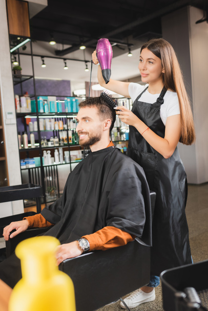 young hairdresser in apron drying hair of smiling man, blurred foreground - Photo, Image