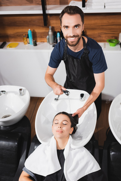 parrucchiere sorridente, barbuto che lava capelli di giovane donna in barbiere - Foto, immagini