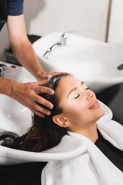 joven mujer sonriendo con los ojos cerrados mientras barbero lavándose el pelo en salón de belleza - Foto, Imagen