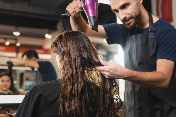 young bearded hairstylist drying hair of woman near blurred mirror reflection - Foto, Bild