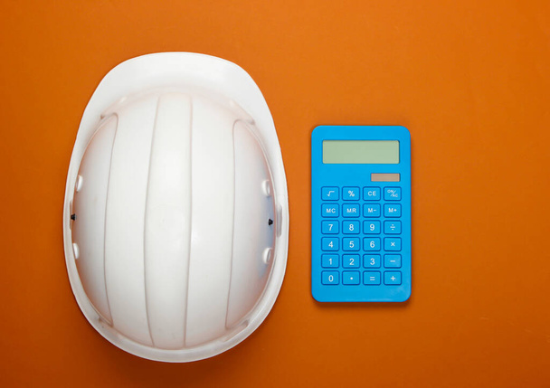 Інженерний будівельний безпечний шолом і калькулятор на помаранчевому фоні. Розрахунок вартості ремонту або будівництва будинку. Вид зверху. Плоский прошарок
 - Фото, зображення