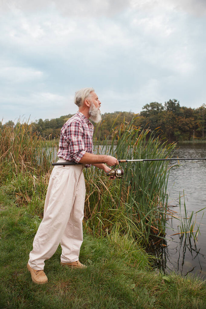 Vertical tiro de longitud completa de un hombre mayor de pesca con una caña en el lago del campo - Foto, Imagen