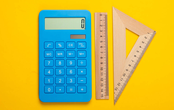 Calculadora y regla de madera, triángulo sobre fondo amarillo. Concepto educativo. Vista superior - Foto, imagen