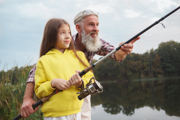 Счастливый старшеклассник с удовольствием обучает свою внучку рыбалке на озере, копировать пространство - Фото, изображение