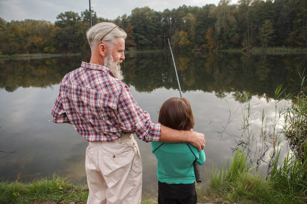 Rückansicht eines älteren Mannes, der seinen Enkel umarmt, während der Junge auf dem See fischt - Foto, Bild