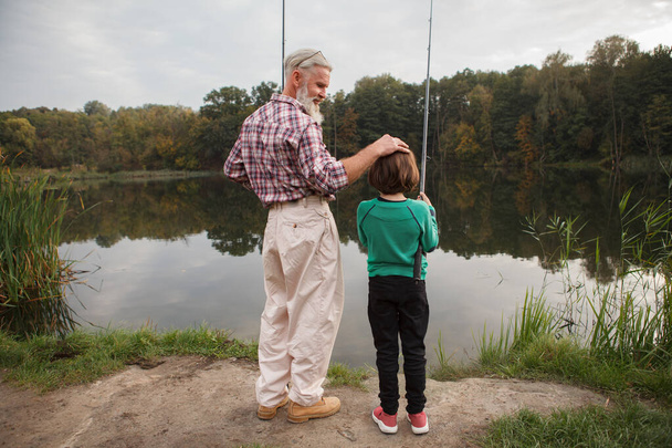 Taustakuva täyspitkä laukaus ylpeä isoisä katsomassa hänen pojanpoikansa kalastaa järvellä - Valokuva, kuva