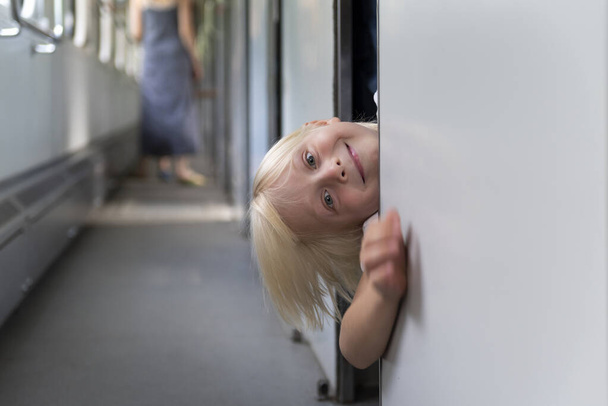A szőke fiú a vonatos kocsiban a fülkéből néz ki. Kisgyermek a vonaton a családi nyaralás. - Fotó, kép