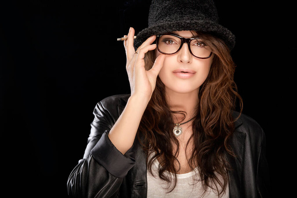 Trendy young woman in leather jacket and matching black hat wearing stylish eyeglasses holding her hand to the frames while smoking a cigarette as she looks at camera with parted lips - Photo, Image