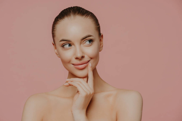 Topless thoughtful lady with clean perfect skin, dark hair, looks aside, undergoes facial trearrments, poses indoor, isolated over rosy background. Cosmetology, skin care and wellness concept - Photo, Image