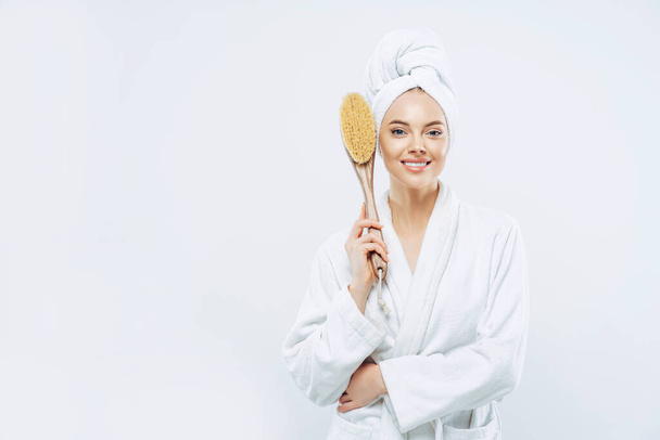 Horizontal shof of charming beauty woman has broad smile, perfect white teeth, dressed in bath robe, holds anti cellulite massage brush, poses against white backgruound, blank space for your promo - Photo, Image