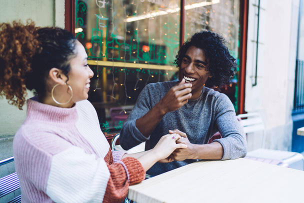 Happy young multiracial couple in casual clothes laughing holding hands and looking to each other while chilling in outdoor cafe in daytime - Фото, изображение