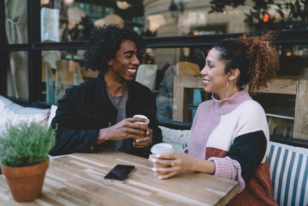 Happy multiracial young man and woman in trendy casual clothes smiling and communicating while having coffee in outdoor street cafe - 写真・画像